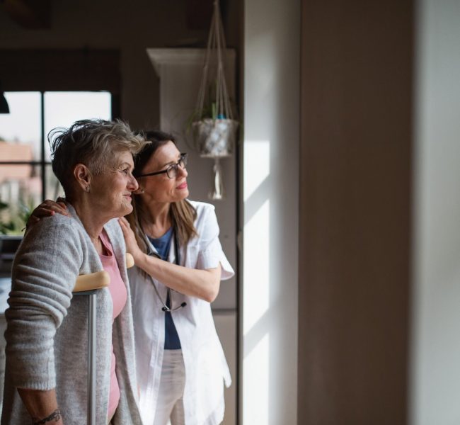 Healthcare worker or caregiver visiting senior woman indoors at home, helping her to walk.